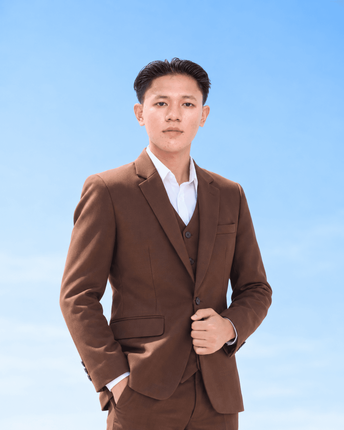 Ari mengenakan setelan jas cokelat, dengan latar langit biru.