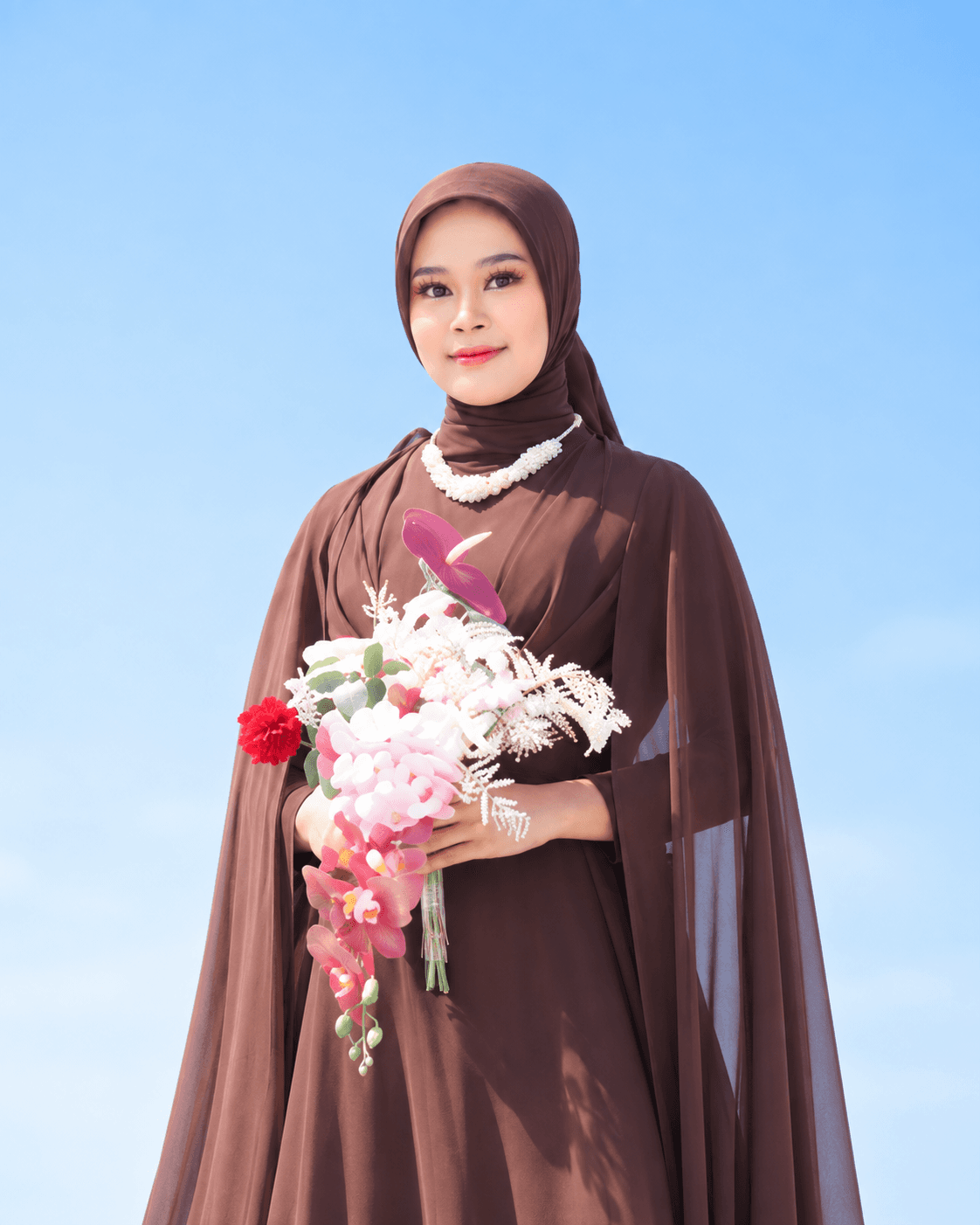 Serly mengenakan gaun dan kerudung cokelat sambil memegang buket bunga, dengan latar langit biru.